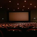 A dimly lit empty cinema theater with rows of red chairs facing a projector screen.